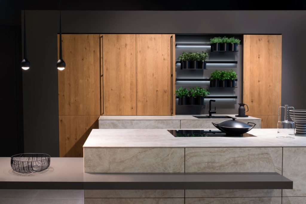 alt="Full frontal view of kitchen with double island with ROCCA fronts, matte dark gray table extension, wall cabinets with MADERO light oak veneers, and inset shelves with hanging plants and hidden LED strip lighting"