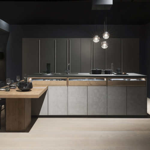alt="frontal view of full showroom kitchen at LEICHT showing concrete doors, madero wood pull outs, and dark, tall cabinets with pearl finish"