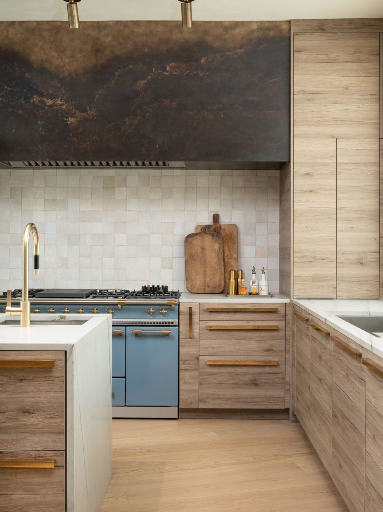 alt="front side kitchen view kitchen cabinets in textured antique oak, wall hood in patinated brass, blue gas range and polished brass hardware"