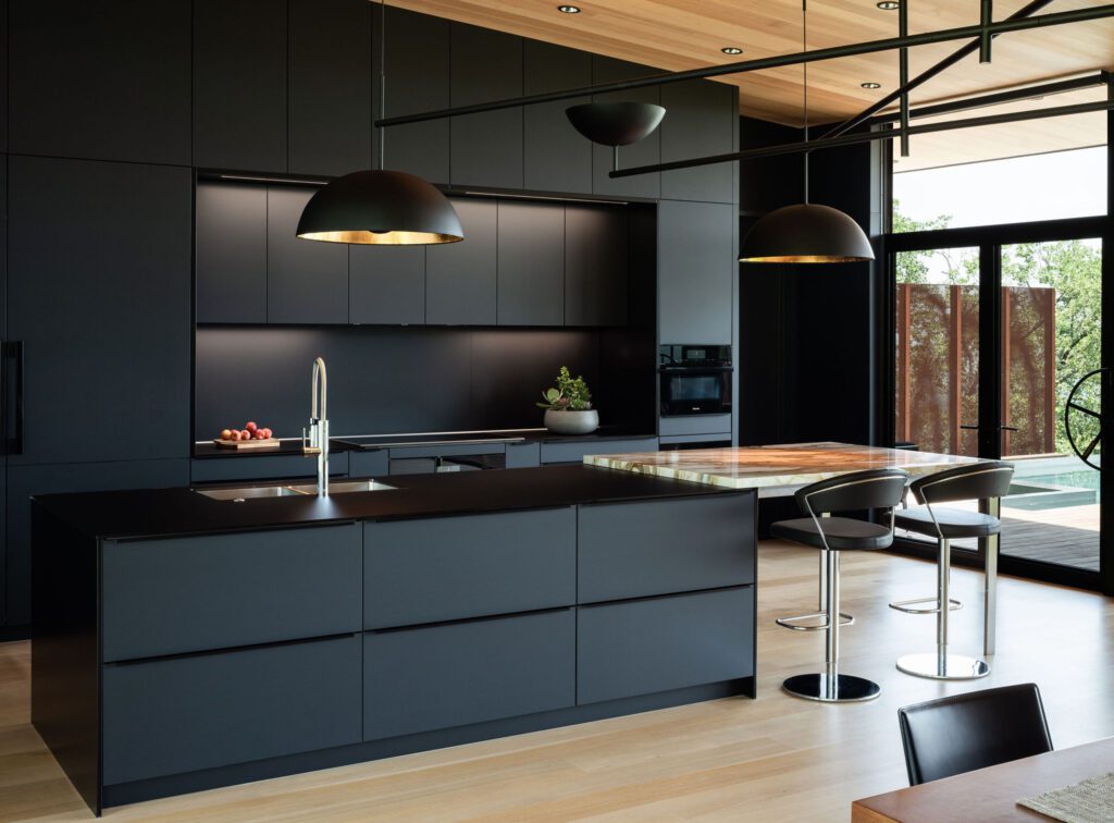 alt=”side kitchen and island view of floor-to-ceiling slate black Fenix cabinetry, color matched matte glass counter top, and contrasting integrated table top”